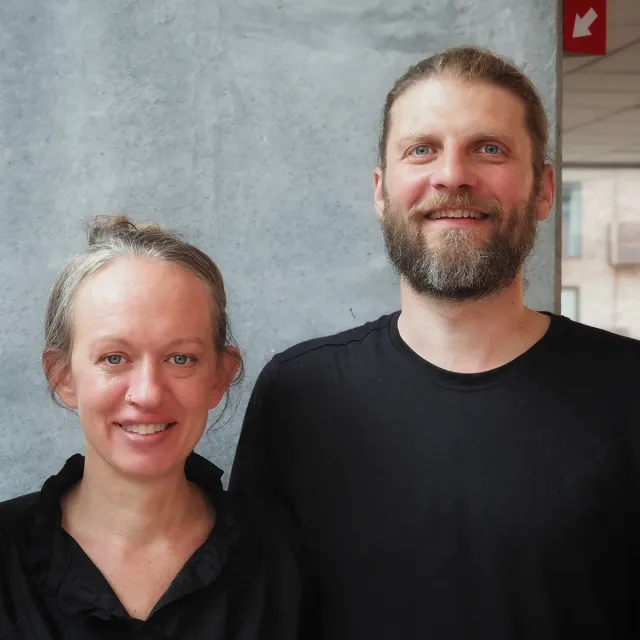 Cathrine og Jonas, yogalærerne bag Øl Yoga, smiler foran en betonvæg